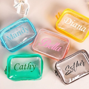 Peut inclure: Cinq trousses de maquillage rectangulaires transparentes de différentes couleurs : bleu, jaune, rose, vert et gris. Chaque trousse a une fermeture éclair et un nom imprimé : Mandy, Diana, Bella, Cathy et Esther. Les trousses sont sur une surface claire.