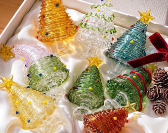 Könnte beinhalten: Eine Sammlung von acht Miniatur-Weihnachtsbaumschmuckstücken aus Glas in verschiedenen Farben wie Bernstein, Transparent, Grün und Rosa, jeweils mit einem gelben Stern gekrönt und mit kleinen farbigen Glaspunkten verziert, in einer weißen Geschenkbox.