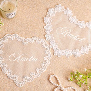 Może przedstawiać: Dwie sercowate koronkowe podkładki z wyhaftowanymi imionami "Amelia" i "Rachel". Podkładki są białe i otoczone kwiatowym koronkowym wykończeniem. Świeca i kwiatowe akcenty znajdują się w tle.