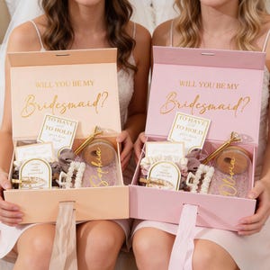 Könnte beinhalten: Zwei offene Geschenkboxen, eine pfirsichfarbene und eine rosa, mit goldenem Text "WILL YOU BE MY Bridesmaid?". Jede Box enthält einen Stift, Haarspangen und andere kleine Geschenke. Die Boxen werden von zwei Personen gehalten.