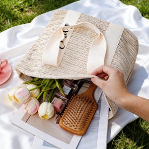 Op de afbeelding: Een beige geweven draagtas met cr&egrave;mekleurige banden, gevuld met items zoals een houten haarborstel, tulpen en parfumflesjes. Een roze kop en schotel staan op een witte doek, met groen gras op de achtergrond.