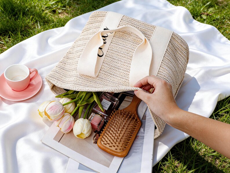 Op de afbeelding: Een beige geweven draagtas met cr&egrave;mekleurige banden, gevuld met items zoals een houten haarborstel, tulpen en parfumflesjes. Een roze kop en schotel staan op een witte doek, met groen gras op de achtergrond.