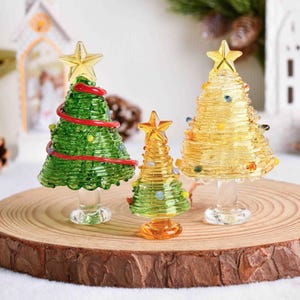May include: Three glass Christmas tree figurines with star toppers. The left tree is green with red garland, the center tree is small and green-yellow, and the right tree is yellow. Each tree features a clear glass pedestal base.