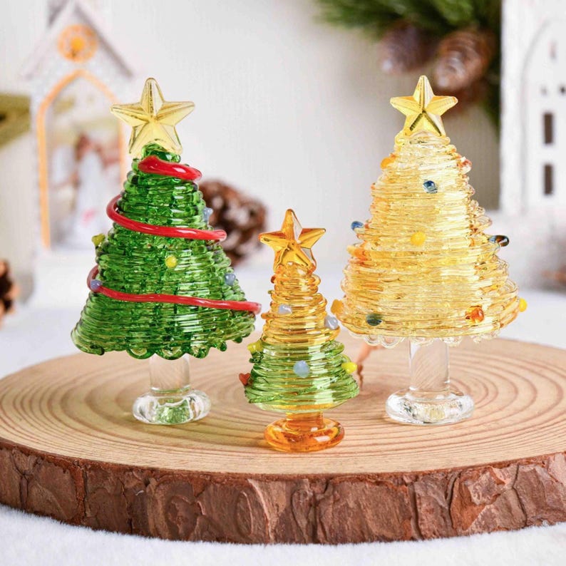 May include: Three glass Christmas tree figurines with star toppers. The left tree is green with red garland, the center tree is small and green-yellow, and the right tree is yellow. Each tree features a clear glass pedestal base.