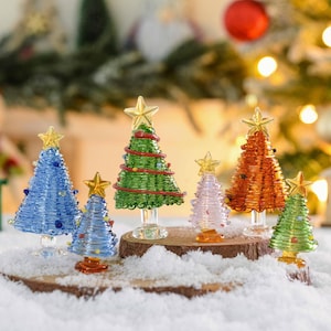 May include: A collection of six miniature glass Christmas trees in various colors including blue, green, pink, and orange. Each tree features a coiled glass design, a clear glass trunk, and a small yellow star topper.