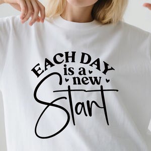 May include: White t-shirt with the black text "EACH DAY is a new Start". The text is in a mix of fonts, with the word "Start" in a large, cursive font. The shirt is plain and simple.