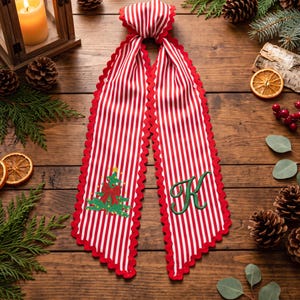 Peut inclure: Un long ruban en tissu à rayures rouges et blanches avec des bords festonnés. Une extrémité présente un sapin de Noël vert brodé avec un nœud rouge, tandis que l'autre extrémité affiche une grande lettre K verte brodée en écriture cursive.