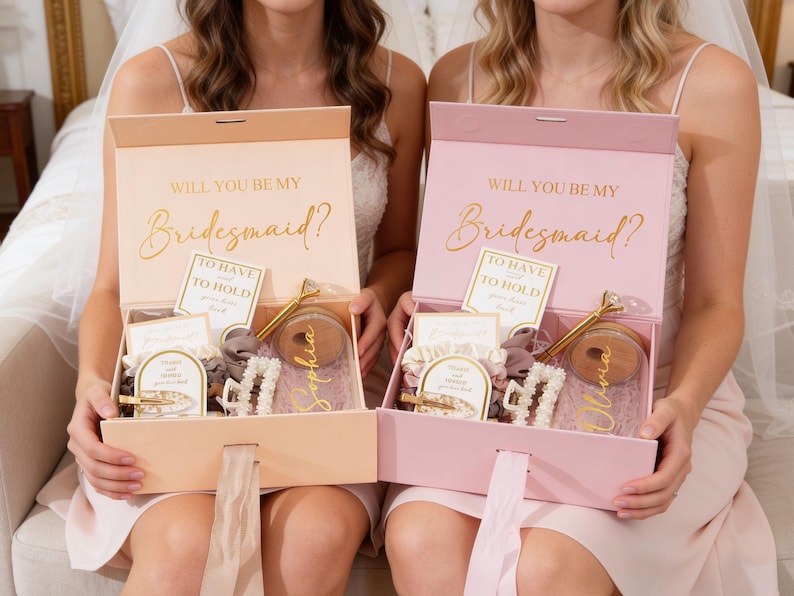 Peut inclure: Deux coffrets cadeaux ouverts, un p&ecirc;che et un rose, avec le texte dor&eacute; "WILL YOU BE MY Bridesmaid ?". Chaque bo&icirc;te contient un stylo, des pinces &agrave; cheveux et d'autres petits cadeaux. Les bo&icirc;tes sont tenues par deux personnes.