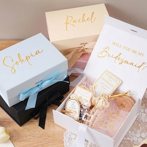 Peut inclure: Plusieurs coffrets cadeaux blancs, noirs et cr&egrave;me avec des lettres dor&eacute;es. Une bo&icirc;te dit "Sohpia" et une autre "Rachel". Une bo&icirc;te blanche ouverte r&eacute;v&egrave;le des cadeaux et le texte "Veux-tu &ecirc;tre ma demoiselle d'honneur ?" avec un ruban.