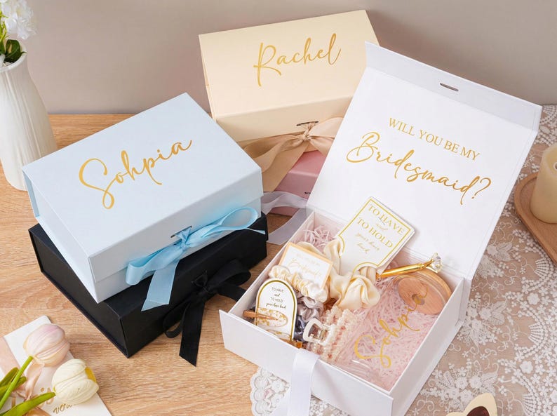 Peut inclure: Plusieurs coffrets cadeaux blancs, noirs et cr&egrave;me avec des lettres dor&eacute;es. Une bo&icirc;te dit "Sohpia" et une autre "Rachel". Une bo&icirc;te blanche ouverte r&eacute;v&egrave;le des cadeaux et le texte "Veux-tu &ecirc;tre ma demoiselle d'honneur ?" avec un ruban.