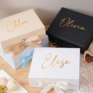 Peut inclure: Quatre bo&icirc;tes cadeaux personnalis&eacute;es en cr&egrave;me, noir, bleu clair et blanc, chacune avec un nom en script dor&eacute;. Les bo&icirc;tes sont nou&eacute;es avec des rubans assortis. Les noms sont Chloe, Olivia et Elise.