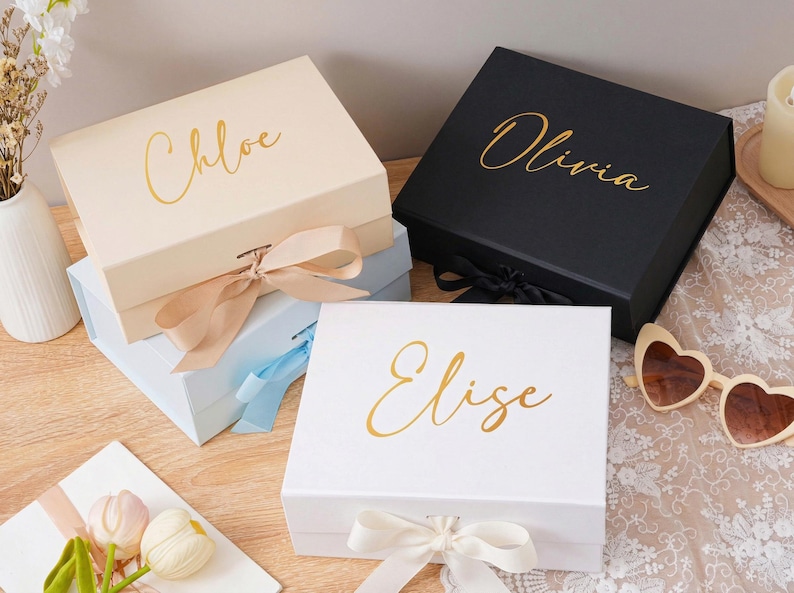 Peut inclure: Quatre bo&icirc;tes cadeaux personnalis&eacute;es en cr&egrave;me, noir, bleu clair et blanc, chacune avec un nom en script dor&eacute;. Les bo&icirc;tes sont nou&eacute;es avec des rubans assortis. Les noms sont Chloe, Olivia et Elise.
