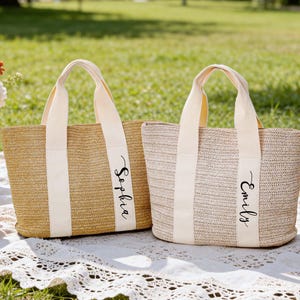 Op de afbeelding: Twee geweven stroken draagtassen met cr&egrave;mekleurige handvatten en verticale strepen. E&eacute;n tas is gemarkeerd met "Sophia" en de andere met "Emily" in zwart schrift. De tassen liggen op een wit kanten picknickkleed met bloemen en champagne.