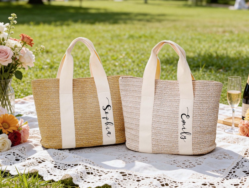 Op de afbeelding: Twee geweven stroken draagtassen met cr&egrave;mekleurige handvatten en verticale strepen. E&eacute;n tas is gemarkeerd met "Sophia" en de andere met "Emily" in zwart schrift. De tassen liggen op een wit kanten picknickkleed met bloemen en champagne.