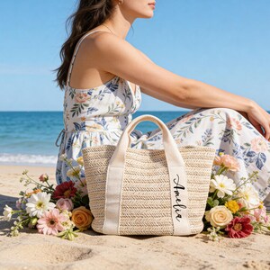 Op de afbeelding: Een beige geweven draagtas met cr&egrave;mekleurige handvatten en een cr&egrave;mekleurige band met de naam "Amelia" in zwart schrift. De tas staat op een zandstrand, omringd door bloemen, met een persoon in een bloemenjurk.