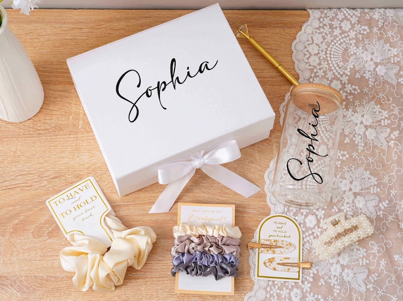 Peut inclure: Une bo&icirc;te cadeau blanche avec le nom "Sophia" en &eacute;criture cursive, un bocal en verre avec un couvercle en bois et le nom "Sophia", et divers accessoires pour cheveux. Les accessoires comprennent des chouchous et des pinces &agrave; cheveux. Les articles sont dispos&eacute;s sur une surface en bois.