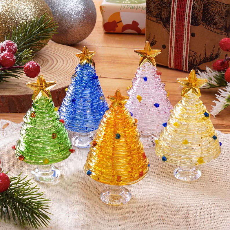 May include: Five miniature glass Christmas trees in green, blue, yellow, clear, and gold colors. Each tree features a coiled glass design, a clear glass pedestal base, and a small gold star topper. Small colored glass beads are embedded throughout.