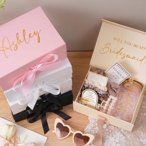 Peut inclure: Bo&icirc;tes cadeaux roses, blanches et noires avec lettrage dor&eacute; et rubans de satin. L'une des bo&icirc;tes est ouverte, r&eacute;v&eacute;lant des accessoires, notamment des chouchous, une pince &agrave; cheveux et un bocal en verre. Le couvercle de la bo&icirc;te ouverte indique "Veux-tu &ecirc;tre ma demoiselle d'honneur ?"