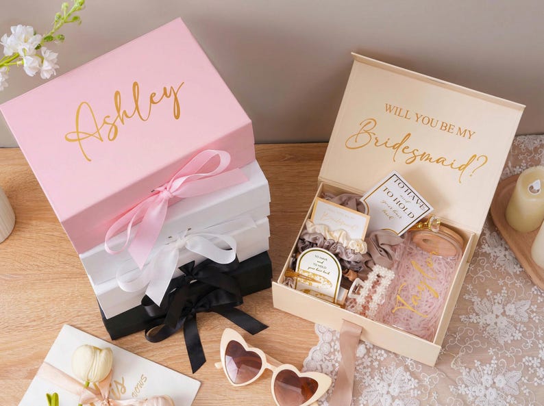 Peut inclure: Bo&icirc;tes cadeaux roses, blanches et noires avec lettrage dor&eacute; et rubans de satin. L'une des bo&icirc;tes est ouverte, r&eacute;v&eacute;lant des accessoires, notamment des chouchous, une pince &agrave; cheveux et un bocal en verre. Le couvercle de la bo&icirc;te ouverte indique "Veux-tu &ecirc;tre ma demoiselle d'honneur ?"