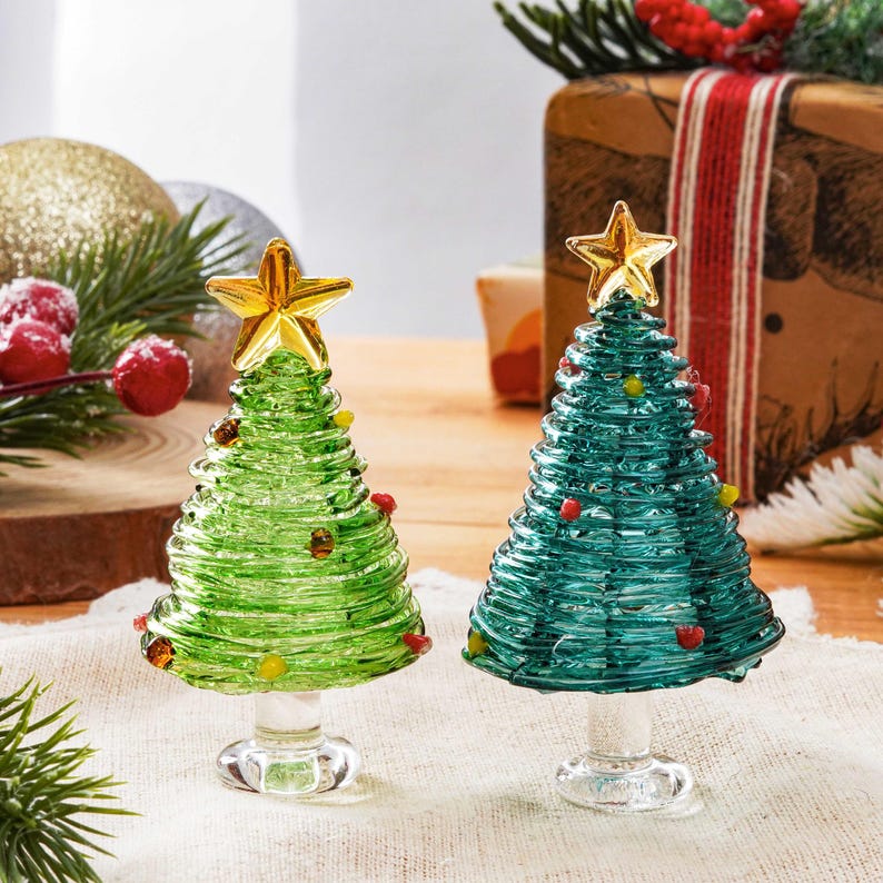 May include: Two glass Christmas tree figurines, one in light green and one in dark teal, each featuring a coiled design, small colorful glass bead accents, and a clear glass base topped with a bright yellow glass star ornament.