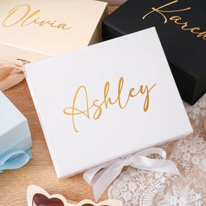 Peut inclure: Plusieurs bo&icirc;tes cadeaux personnalis&eacute;es de diff&eacute;rentes couleurs, dont blanc, noir et bleu clair. Chaque bo&icirc;te porte un nom en lettres dor&eacute;es, comme "Ashley" et "Karen", et est nou&eacute;e avec un ruban assorti. Les bo&icirc;tes sont dispos&eacute;es sur une surface en bois.