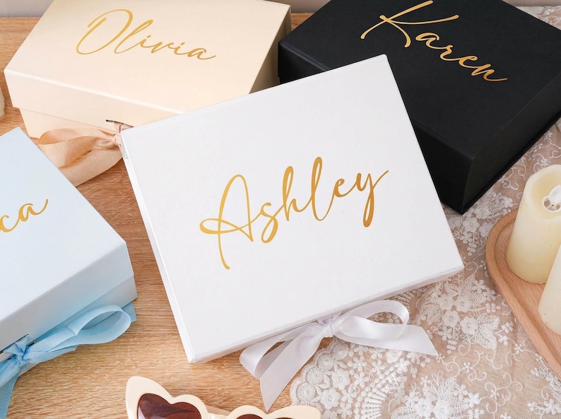 Peut inclure: Plusieurs bo&icirc;tes cadeaux personnalis&eacute;es de diff&eacute;rentes couleurs, dont blanc, noir et bleu clair. Chaque bo&icirc;te porte un nom en lettres dor&eacute;es, comme "Ashley" et "Karen", et est nou&eacute;e avec un ruban assorti. Les bo&icirc;tes sont dispos&eacute;es sur une surface en bois.