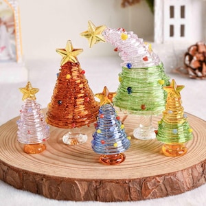 May include: A collection of five glass Christmas tree figurines in various colors including amber, light blue, green, and clear. Each tree features a coiled glass design, small colorful glass bead ornaments, and a gold-toned star topper.