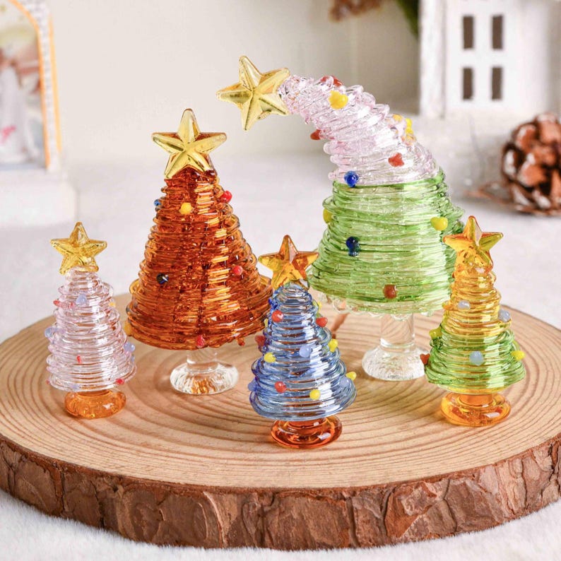 May include: A collection of five glass Christmas tree figurines in various colors including amber, light blue, green, and clear. Each tree features a coiled glass design, small colorful glass bead ornaments, and a gold-toned star topper.