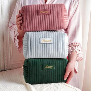 Può includere: Tre trousse per cosmetici in velluto a coste rosa, azzurro e verde, impilate l'una sull'altra. Ogni borsa ha un'etichetta rettangolare con un nome ricamato in oro. Le borse hanno una chiusura a cerniera.