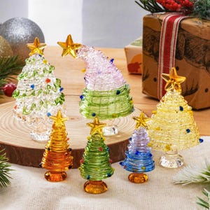 May include: A collection of seven glass Christmas tree figurines in various colors including clear, green, yellow, orange, and blue. Each tree features a coiled glass design, small colorful glass bead ornaments, and a yellow star topper.