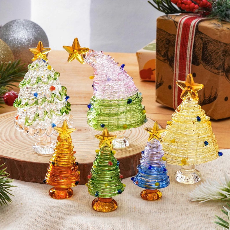 May include: A collection of seven glass Christmas tree figurines in various colors including clear, green, yellow, orange, and blue. Each tree features a coiled glass design, small colorful glass bead ornaments, and a yellow star topper.