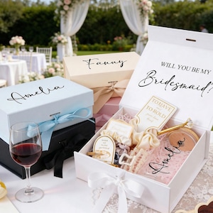 Peut inclure: Plusieurs coffrets cadeaux, dont une bo&icirc;te blanche avec le texte "Will You Be My Bridesmaid ?" et une bo&icirc;te bleu clair avec le nom "Amelia". Les bo&icirc;tes sont nou&eacute;es avec des rubans et contiennent divers cadeaux, pos&eacute;s sur une table avec un verre de vin rouge.