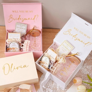 Peut inclure: Coffrets cadeaux roses et blancs avec l'inscription dor&eacute;e "Will You Be My Bridesmaid ?" et "Olivia". Les bo&icirc;tes contiennent divers cadeaux, dont un bocal en verre, des chouchous et des cartes. Les bo&icirc;tes sont ouvertes, r&eacute;v&eacute;lant le contenu.