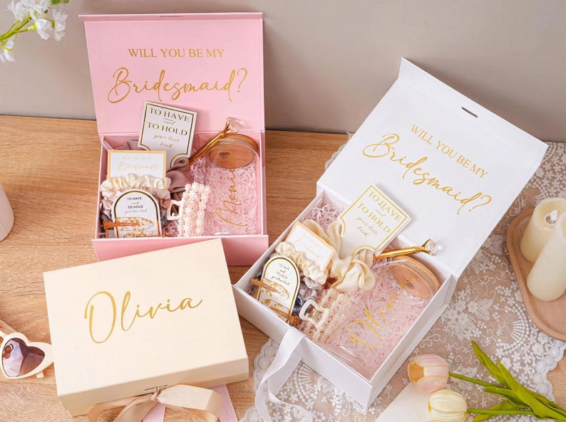 Peut inclure: Coffrets cadeaux roses et blancs avec l'inscription dor&eacute;e "Will You Be My Bridesmaid ?" et "Olivia". Les bo&icirc;tes contiennent divers cadeaux, dont un bocal en verre, des chouchous et des cartes. Les bo&icirc;tes sont ouvertes, r&eacute;v&eacute;lant le contenu.