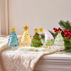 May include: Five miniature glass Christmas trees in various colors including light blue, yellow, green, pink, and clear. Each tree features a coiled glass design, small colorful glass bead accents, and a gold-toned star topper.