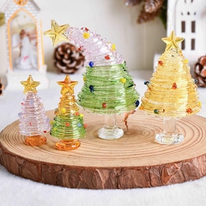 May include: Four glass Christmas tree figurines with coiled designs and star toppers. Two trees are yellow, one is green with a curved top, and one is clear with a pink tint. Each tree features small colorful glass beads and a clear pedestal base.