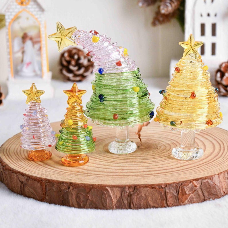 May include: Four glass Christmas tree figurines with coiled designs and star toppers. Two trees are yellow, one is green with a curved top, and one is clear with a pink tint. Each tree features small colorful glass beads and a clear pedestal base.