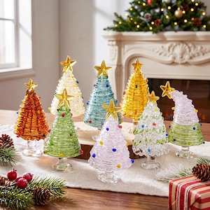 May include: A collection of eight small glass Christmas tree figurines in various colors including red, green, blue, yellow, and clear, each topped with a gold star and decorated with small colored glass dots on a clear glass pedestal base.