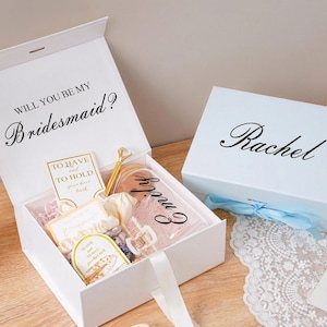 Peut inclure: Bo&icirc;tes cadeaux blanches, l'une ouverte avec le texte "Will you be my Bridesmaid ?", l'autre ferm&eacute;e avec le texte "Rachel". La bo&icirc;te ouverte contient des cadeaux, dont un accessoire pour cheveux et une carte. Un ruban bleu clair orne la bo&icirc;te ferm&eacute;e.