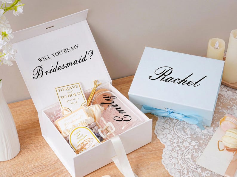 Peut inclure: Bo&icirc;tes cadeaux blanches, l'une ouverte avec le texte "Will you be my Bridesmaid ?", l'autre ferm&eacute;e avec le texte "Rachel". La bo&icirc;te ouverte contient des cadeaux, dont un accessoire pour cheveux et une carte. Un ruban bleu clair orne la bo&icirc;te ferm&eacute;e.