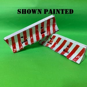 May include: Two miniature traffic barriers featuring a white base with vertical red stripes. The barriers have a sloped top edge and two circular holes on the front face. The text SHOWN PAINTED is printed in black above the two objects.