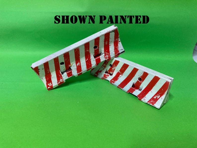May include: Two miniature traffic barriers featuring a white base with vertical red stripes. The barriers have a sloped top edge and two circular holes on the front face. The text SHOWN PAINTED is printed in black above the two objects.