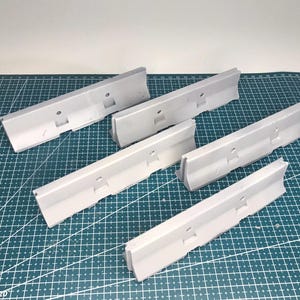 May include: A set of five light gray plastic jersey barriers, each featuring a sloped profile, a flat base, and a small circular hole centered along the top edge. These miniature road dividers are arranged in a scattered formation.