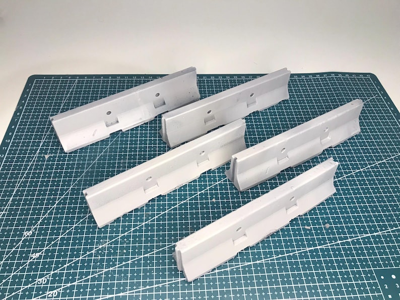 May include: A set of five light gray plastic jersey barriers, each featuring a sloped profile, a flat base, and a small circular hole centered along the top edge. These miniature road dividers are arranged in a scattered formation.