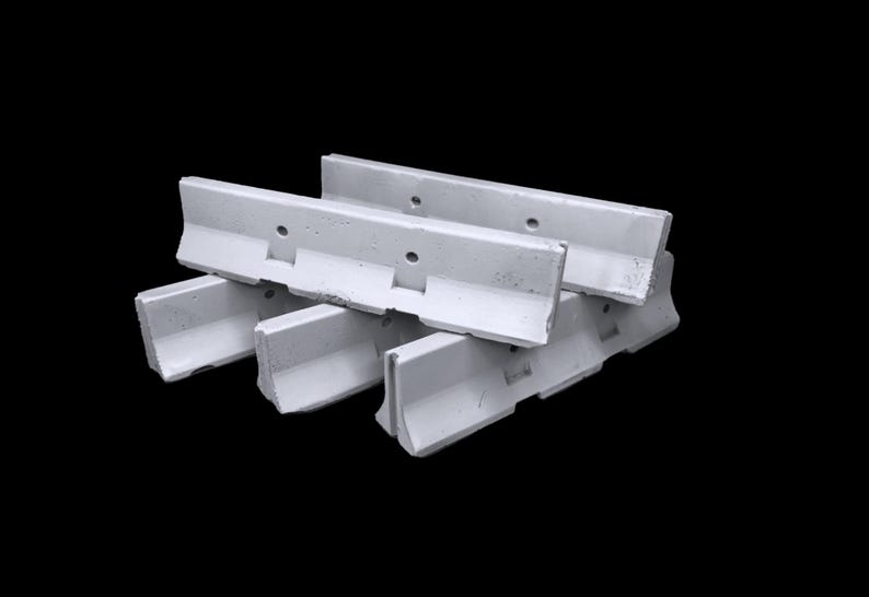 May include: A set of three light gray concrete jersey barriers stacked in a cluster. Each barrier features a distinct L-shaped profile with a wide base, a vertical wall section, and two circular holes positioned along the upper edge of the wall.