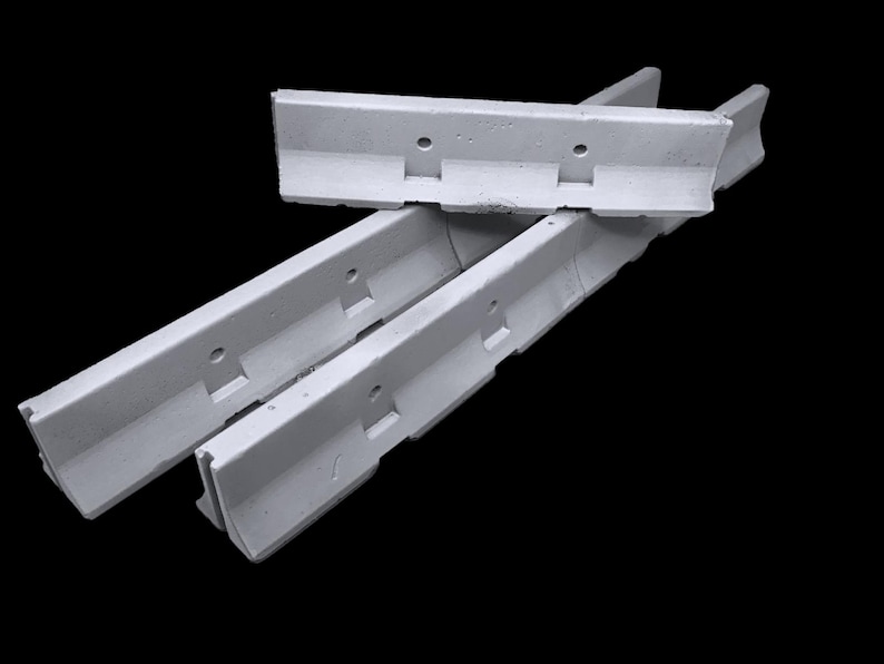 May include: Three light gray concrete barriers with a linear, elongated shape. Each barrier features a distinct L-shaped profile with a flat base, a vertical wall section, and two circular holes positioned along the upper edge of the wall.