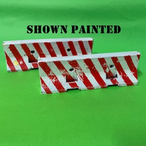 May include: Two miniature concrete-style traffic barriers featuring diagonal red and white hazard stripes. The barriers have a weathered, distressed finish with visible paint chips. The text SHOWN PAINTED is printed in black above the objects.