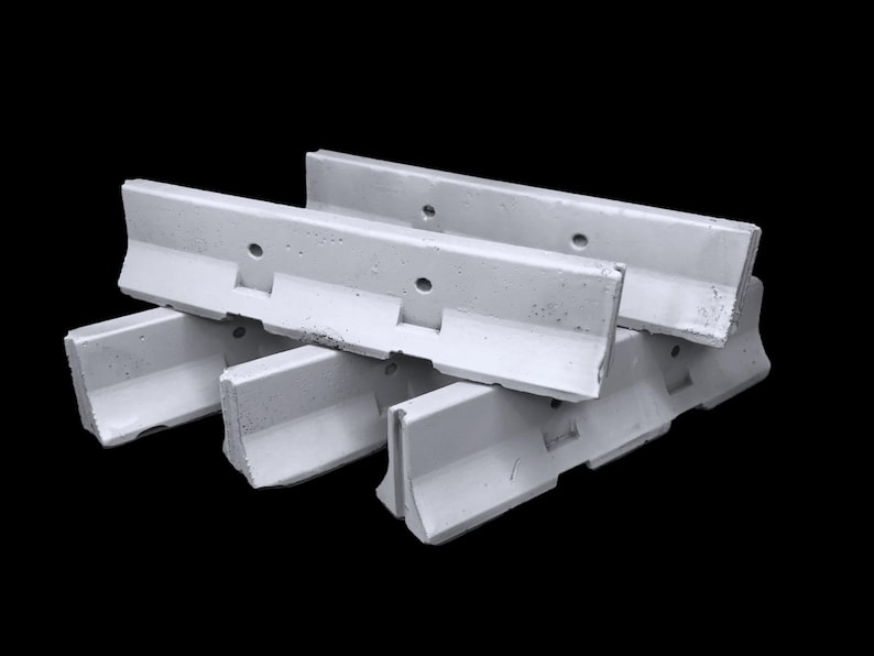 May include: A stack of three white concrete traffic barriers featuring a low-profile design with a wide base, a sloped front face, and a vertical top section. Each barrier includes two circular holes positioned horizontally along the upper edge.