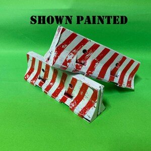 May include: Two miniature road barriers featuring a white base with diagonal red stripes and a weathered, distressed paint finish. The text SHOWN PAINTED is printed in black capital letters above the two barriers, which have a hollow design.