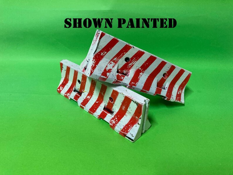May include: Two miniature road barriers featuring a white base with diagonal red stripes and a weathered, distressed paint finish. The text SHOWN PAINTED is printed in black capital letters above the two barriers, which have a hollow design.